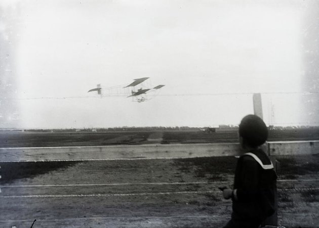 Farman gép a levegőben. Az előtérben a versenyek idejére épített ideiglenes kerítés egy kíváncsiskodó gyermekkel, a háttérben a pálya jelzőtornya és egy lovaskocsi (kép forrása: Fortepan/ Magyar Műszaki és Közlekedési Múzeum / Archívum / Negatívtár / Tas Gyula gyűjteménye)